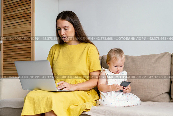 Unplugging mother after work. Child using mobile phone while mom working remotly at home. Стоковое фото, фотограф Natalia Gdovskaia / Фотобанк Лори