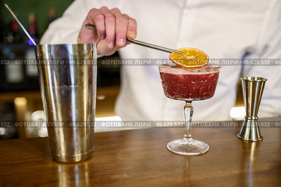 Unrecognizable male barkeeper putting dried slice of orange on top of cold fruit cocktail placed on counter in pub. Crop barman garnishing cocktail with citrus slice. Стоковое фото, агентство Ingram Publishing / Фотобанк Лори
