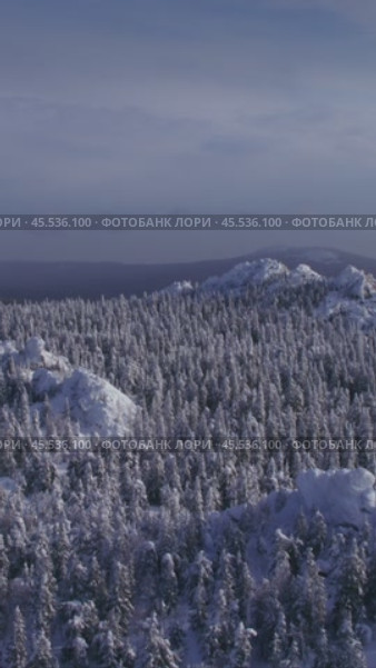 Вертикальное видео: вид с воздуха на хребты Южного Урала зимой. Заснеженные горные пики возвышаются среди леса. Vertical video: aerial view of the snow-covered ridges of the Southern Urals during winter. Mountain peaks rise among the forests. Редакционное видео, видеограф Валерий Тесаков / Фотобанк Лори