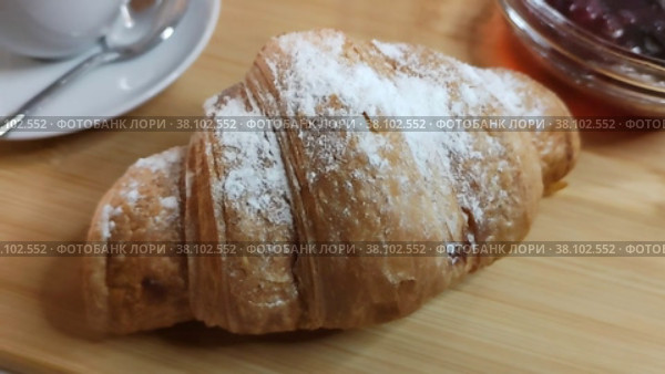 Вкусный хрустящий круассан  посыпанный сахарной пудрой на деревянной доске крупным планом. Традиционное кондитерское изделие французской кухни. Круассан с белой посыпкой, варенье и кофе для континентального завтрака. Delicious crispy croissant sprinkled with powdered sugar on wooden board close-up. Traditional confection of French cuisine. Croissant with white sprinkles, jam and coffee for continental breakfast. Стоковое видео, видеограф Ирина / Фотобанк Лори