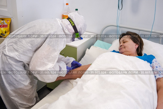 Врач в защитном костюме от COVID-19 берёт кровь на анализы у больной в палате инфекционного отделения, в красной зоне медицинского центра ​помощи и спасения. Богучаны, Красноярский край, Россия (2021 год). Редакционное фото, фотограф Светлана Попова / Фотобанк Лори