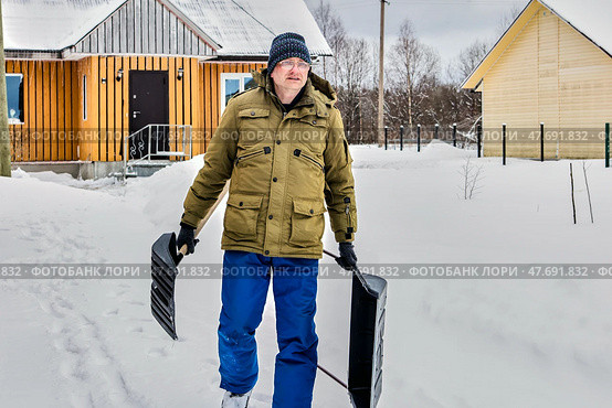 Walking through snowy yard mature man in warm jacket holds large two handed snow pusher in one hand and smaller snow shovel in other. Стоковое фото, фотограф Владимир Григорьев / Фотобанк Лори