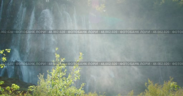Waterfall mist rises high in forest air as water cascades down rocky green cliff face. Стоковое видео, видеограф Ольга Сухарева / Фотобанк Лори