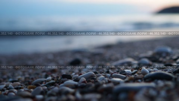 Waves gently lap at rocky shore during stunning sunset over ocean. Blue sunlight reflects off waves as they roll onto shore, creating tranquil atmosphere at sunset by sea. Focus on pebbles and stones. Стоковое видео, видеограф Юлия Петровская / Фотобанк Лори