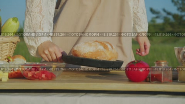 Woman, clad in rustic dress and apron, carries tray of freshly baked bread to table during summer harvest of vegetables. Стоковое видео, видеограф Strunov David / Фотобанк Лори