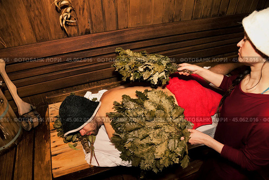 Woman in bath uniform doing whipped with broom steamed human male body in hot steam sauna, russian bath. Male lying relaxing on wood bench. Wellness, self-care, healthy concept. Copy ad text space. Стоковое фото, фотограф Aleksandr Golubev / Фотобанк Лори