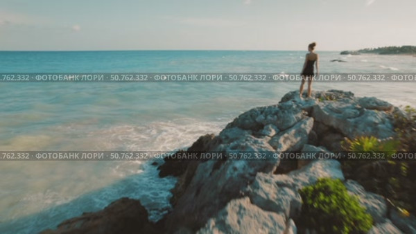 Woman in black dress stands alone on rocky point looking out over calm ocean small waves and sandy seabed in Tulum, Mexico. Стоковое видео, видеограф Igor Tichonow / Фотобанк Лори