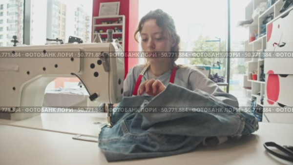 Woman is crafting jeans by sewing them on industrial machine. Measuring tape is draped around her neck as she carefully guides fabric. Стоковое видео, видеограф Strunov David / Фотобанк Лори