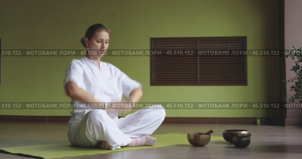 Woman practicing kundalini yoga, doing breathing exercise, clasping hands together, lowering them sharply on exhalation, sitting in white comfortable clothes on mat in lotus position in room. Стоковое видео, видеограф Сергей Чайко / Фотобанк Лори