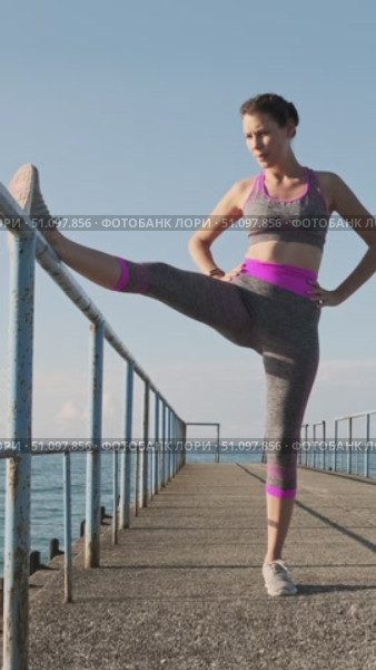 Woman stretching leg on metal railing while standing on concrete pier by the sea under morning sunlight, front view fitness routine improving flexibility, balance and active healthy coastal lifestyle. Стоковое видео, видеограф Сергей Тимофеев / Фотобанк Лори