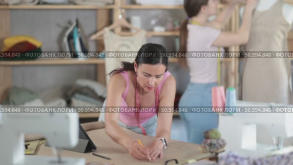 Woman tailor draws dress diagram scheme on paper. Companion worker in background with mannequin. Стоковое видео, видеограф Яков Филимонов / Фотобанк Лори