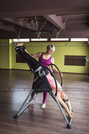 Woman trainer giving inversion therapy to student, hanging upside down on inversion table in room. Стоковое фото, фотограф Сергей Чайко / Фотобанк Лори