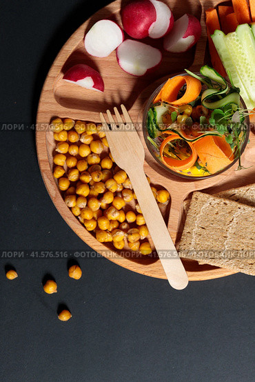 Wooden menagerie with healthy snacks, pieces of fresh vegetables, crispy chickpeas, grain loaves. Close-up on a black background. Стоковое фото, фотограф Bogdanova Mariia / Фотобанк Лори