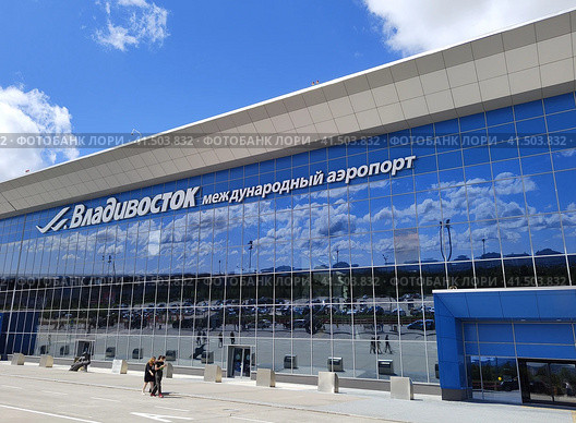 Ярко-голубое небо с белыми облаками отражается в зеркальном фасаде здания. Терминал международного аэропорта. Кневичи. Владивосток, Россия, 26 августа 2023. Редакционное фото, фотограф Ирина / Фотобанк Лори