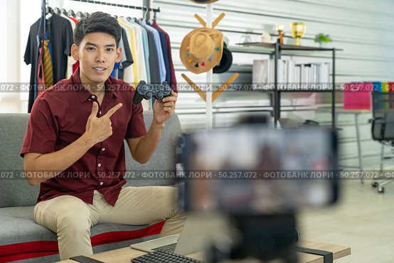 Young asian male online blogger recording live vlog video for review joystick for gaming IT product goods. Online influcencer on social media concept. Focus on mobile phone. Стоковое фото, агентство Ingram Publishing / Фотобанк Лори