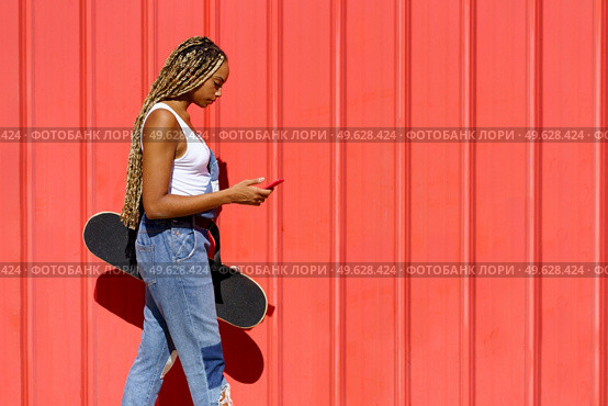 Young black girl with coloured braids walking on an urban red background while consulting her smartphone.. Black girl walking on an urban red background while consulting her smartphone. Стоковое фото, агентство Ingram Publishing / Фотобанк Лори