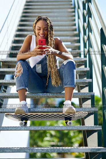 Young black woman, with coloured braids, watching something very funny on her smartphone. Girl with a skateboard.. Black woman, with coloured braids, watching something funny on her smartphone. Стоковое фото, агентство Ingram Publishing / Фотобанк Лори