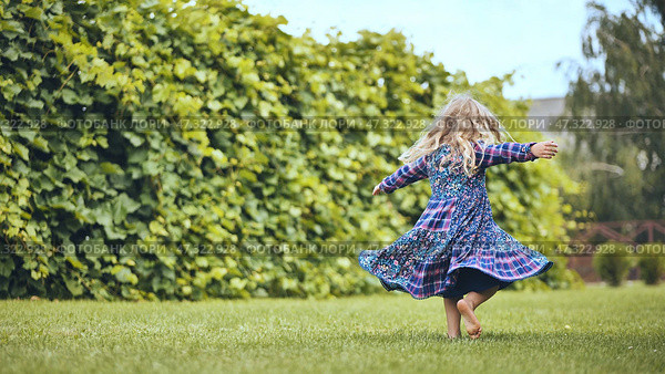 Young blonde girl spinning playfully in white sundress, dancing barefoot on lush green lawn near garden hedgerow, radiating childhood joy. Стоковое фото, фотограф Mikhail Davidovich / Фотобанк Лори
