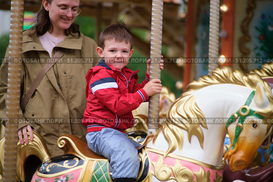 Young boy in red jacket enjoys a ride on a colorful carousel horse, accompanied by smiling woman during a fun outdoor outing. A child aged 3 years. Стоковое фото, фотограф Андрей Журавлев / Фотобанк Лори