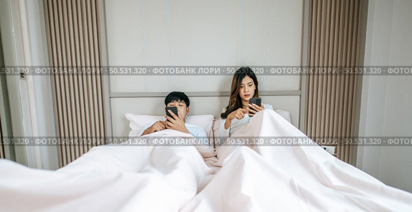 Young Couple lying on bed in bedroom, They are enjoy with social media networks addicts surf the internet on mobile phones ignoring each other, family addicted smartphone concept. Стоковое фото, агентство Ingram Publishing / Фотобанк Лори