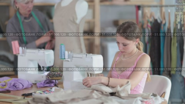 Young dressmaker works on a sewing machine against the background of a mature worker who checks a sample of a dress on a mannequin. Dressmakers work in the sewing industry. Стоковое видео, видеограф Яков Филимонов / Фотобанк Лори