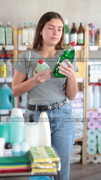 Young female customer in home essentials store holding two bottles of dishwashing liquid, carefully comparing product features before making decision. Стоковое видео, видеограф Яков Филимонов / Фотобанк Лори