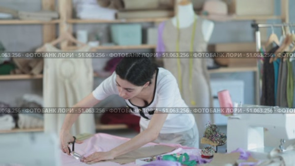 Young female fashion designer stands near a table in a sewing workshop and cuts fabric, checking the paper template. Process of tailoring clothes. Стоковое видео, видеограф Яков Филимонов / Фотобанк Лори