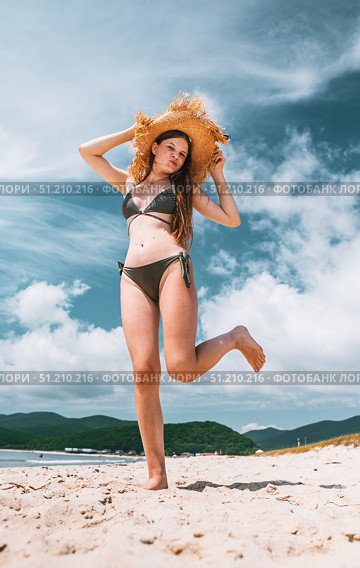 Young girl in bikini and straw hat posing playfully on sandy beach under blue sky. Concept of summer fashion, beach lifestyle, holiday vibes, and freedom mood. Стоковое фото, фотограф Енваев Андрей / Фотобанк Лори