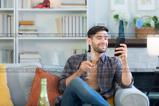 Young man drinking champagne and using smartphone at home in the living room. Стоковое фото, агентство Ingram Publishing / Фотобанк Лори