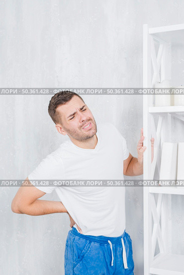 young man standing against concrete wall touching his back grimacing face with pain. Стоковое фото, агентство Ingram Publishing / Фотобанк Лори