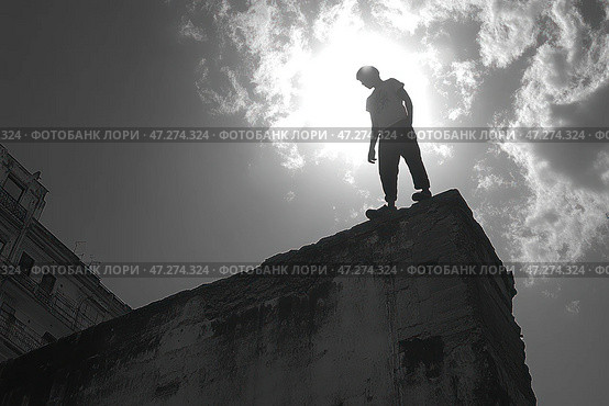 Young man standing on the edge of an abandoned building, silhouetted against a bright sun surrounded by clouds. The shot is taken from below, enhancing the sense of height and the contrast between the man and the sky. The atmosphere is tense, ambiguous, perhaps symbolizing a search for meaning, risk, or overcoming fears. The buildings in the background emphasize the urban nature of the scene, adding a sense of loneliness or contemplation of the future. Стоковое AI-изображение, AI-художник Анна Кривицкая / Фотобанк Лори