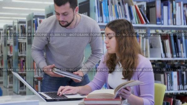 Young positive interested couple of students spending time together in library, reading books and browsing educational websites on laptop. Стоковое видео, видеограф Яков Филимонов / Фотобанк Лори