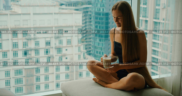 Young smiling woman drinking delicious coffee near window with big city skyscrapers outside. Relaxed girl enjoys morning cappuccino on weekend in the metropolis. Slow motion close-up footage. Стоковое фото, фотограф Ольга Сухарева / Фотобанк Лори
