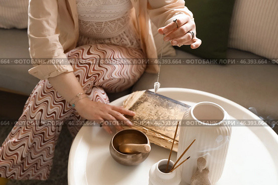 Young witch with pendulum and spell book on table at home, closeup. Стоковое фото, фотограф Pixel_shot / Фотобанк Лори