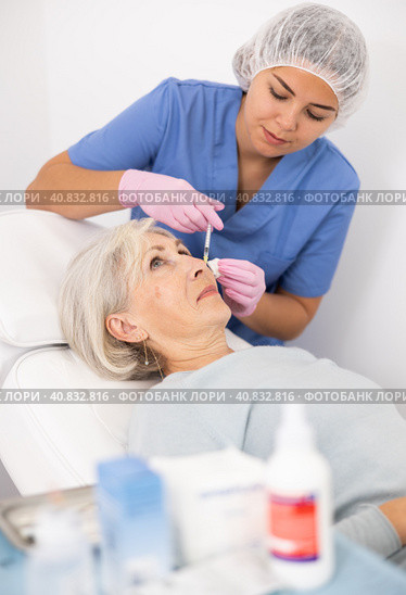 Young woman beautician performing procedure of injection biorevitalization to mature woman for alignment of facial asymmetry in esthetic clinic. Стоковое фото, фотограф Яков Филимонов / Фотобанк Лори