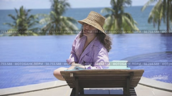 Young woman sits on a lounge chair by a pool at eco beach resort hotel, enjoying the ocean view and palm trees and talking something. Relaxation and tropical vacation vibes. Summer concept. Стоковое видео, видеограф Ирина Михайличенко / Фотобанк Лори