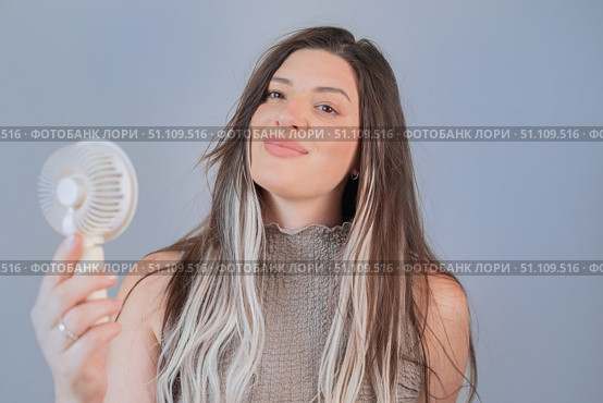 Young woman smiling while holding a portable mini fan, enjoying a cool breeze on a hot summer day. Стоковое фото, фотограф Svetlana Golovco / Фотобанк Лори