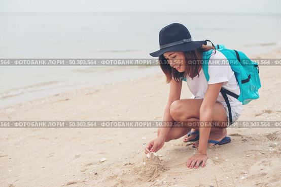 Young woman with backpack sitting on beach and try to make a castle sand. Стоковое фото, агентство Ingram Publishing / Фотобанк Лори