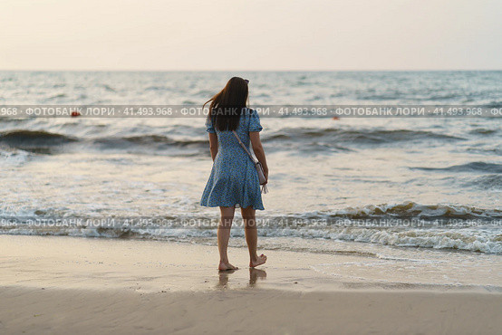 Zelenogradsk, Russia - August 24, 2022: Photography of the amazing Baltic sea. Waves going onto sandy beach. Beautiful young woman standing on the beach. The wind blows her hair and light dress. Concept of beauty in nature. Back, rear view. Редакционное фото, фотограф YuryKara / Фотобанк Лори