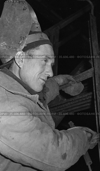 Зрелый мужчина работающий сварщиком с защитной маской на голове, человек в рабочей одежде и перчатках спускаются по металлической лестнице, возведение Чульманской электростанции, Якутия, 1960-е. Редакционное фото, фотограф Варфоломей Тетерин / Фотобанк Лори